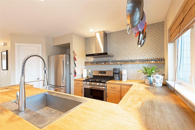 kitchen with sink, wall chimney exhaust hood, tasteful backsplash, wooden counters, and appliances with stainless steel finishes