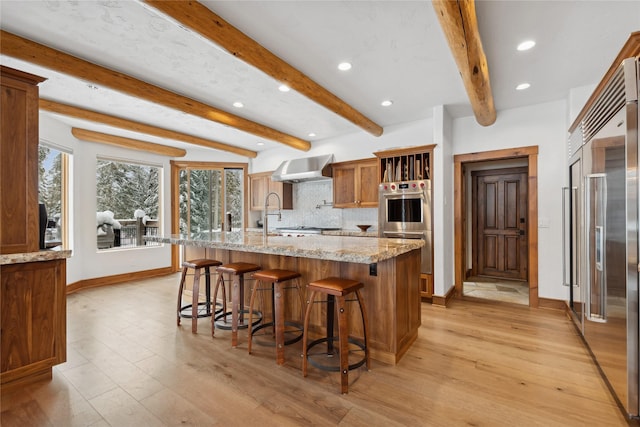 kitchen featuring wall chimney range hood, stainless steel appliances, an island with sink, decorative backsplash, and light stone counters