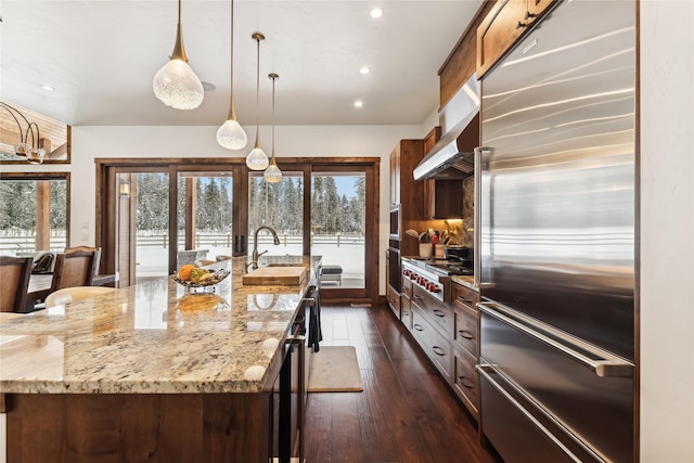 kitchen featuring appliances with stainless steel finishes, sink, light stone countertops, a large island with sink, and wall chimney range hood