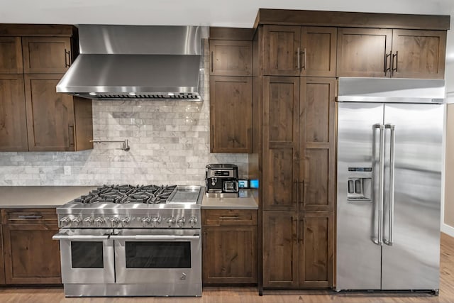 kitchen with high end appliances, decorative backsplash, wall chimney exhaust hood, and dark brown cabinets