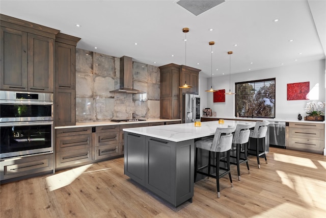 kitchen featuring pendant lighting, wall chimney range hood, a large island, appliances with stainless steel finishes, and light hardwood / wood-style floors