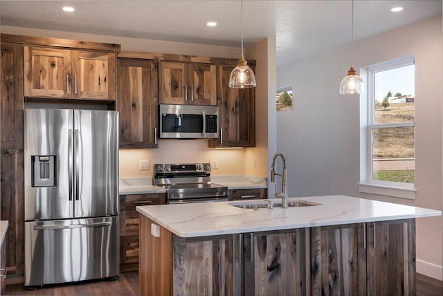 kitchen with pendant lighting, sink, light stone countertops, an island with sink, and appliances with stainless steel finishes