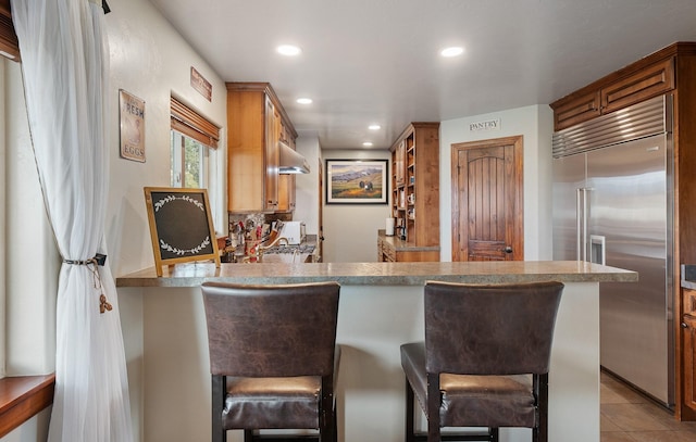 kitchen with exhaust hood, kitchen peninsula, light tile patterned flooring, a kitchen breakfast bar, and stainless steel built in refrigerator