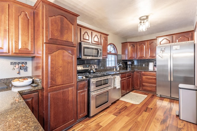 kitchen with stainless steel appliances, dark stone counters, sink, backsplash, and light hardwood / wood-style flooring