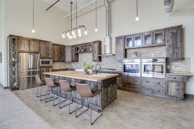 kitchen featuring butcher block counters, wall chimney range hood, stainless steel appliances, dark brown cabinets, and a center island with sink