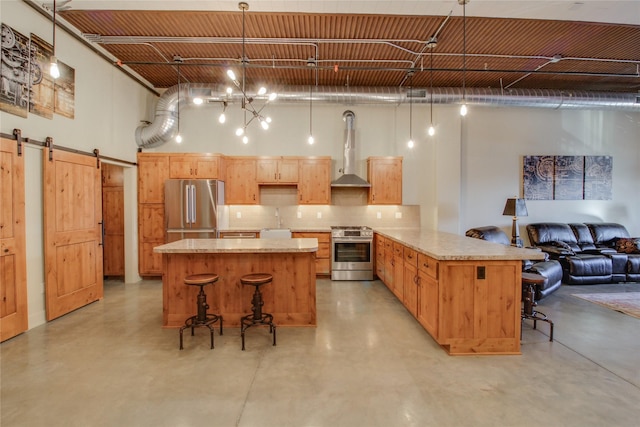 kitchen featuring wall chimney range hood, decorative light fixtures, a barn door, and appliances with stainless steel finishes