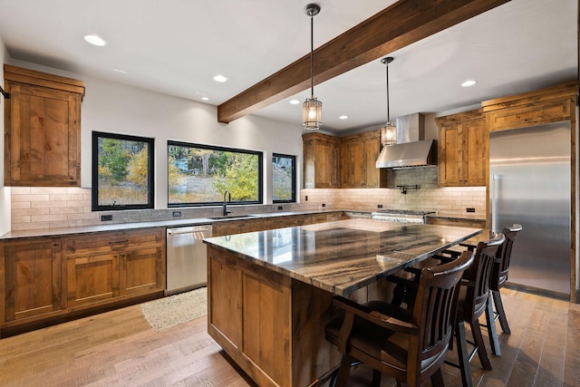 kitchen with a center island, wall chimney exhaust hood, stainless steel appliances, dark stone counters, and beam ceiling