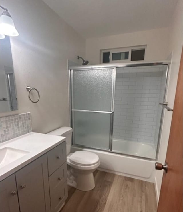 full bathroom featuring bath / shower combo with glass door, backsplash, hardwood / wood-style flooring, vanity, and toilet