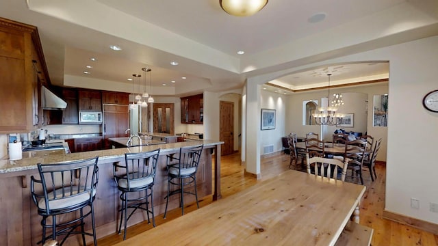 kitchen with light wood-type flooring, stainless steel microwave, a tray ceiling, light stone countertops, and exhaust hood