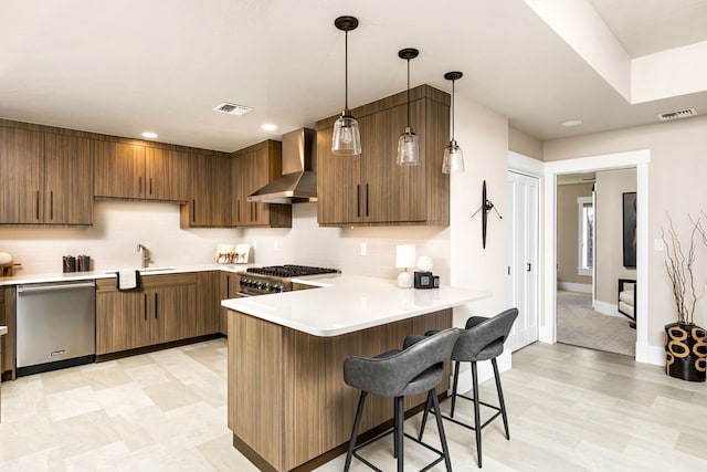 kitchen with appliances with stainless steel finishes, hanging light fixtures, a kitchen breakfast bar, kitchen peninsula, and wall chimney exhaust hood