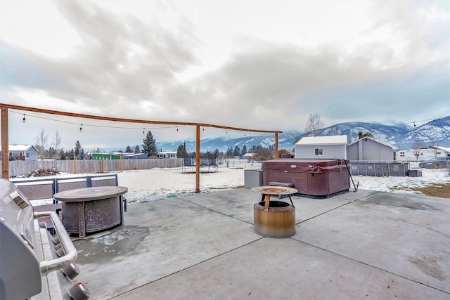 snow covered patio with a hot tub, a mountain view, a trampoline, and an outdoor fire pit