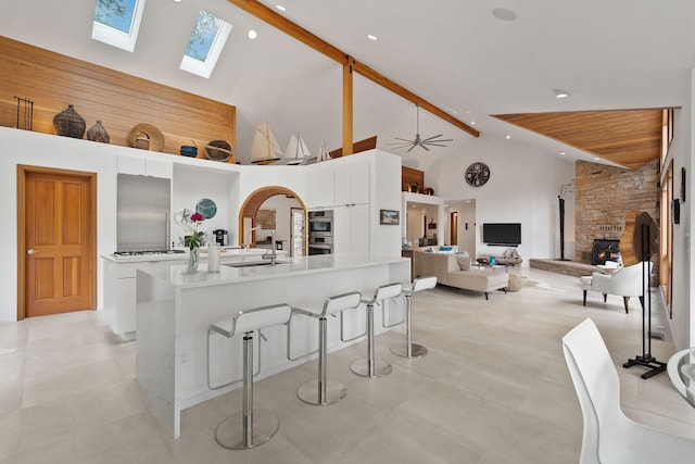 kitchen featuring a stone fireplace, high vaulted ceiling, a center island with sink, a kitchen breakfast bar, and white cabinets