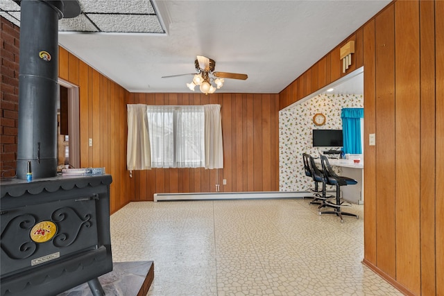 office area with a baseboard heating unit, ceiling fan, a wood stove, and wooden walls