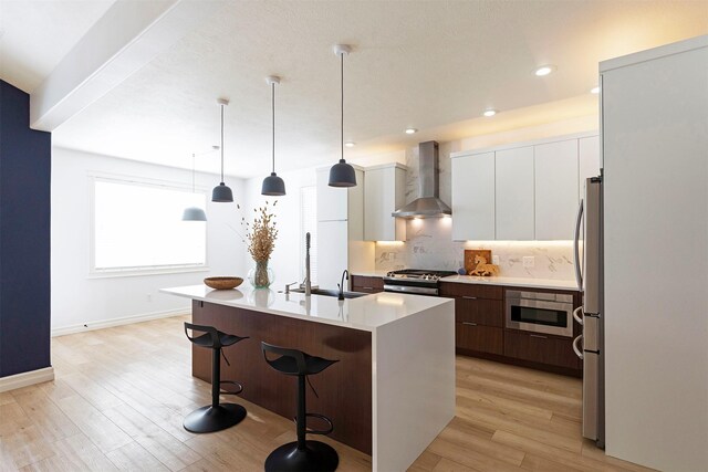 kitchen featuring white cabinetry, a center island with sink, pendant lighting, stainless steel appliances, and wall chimney range hood