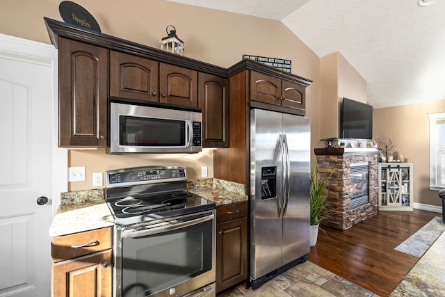 kitchen featuring light stone counters, vaulted ceiling, dark brown cabinets, stainless steel appliances, and a fireplace