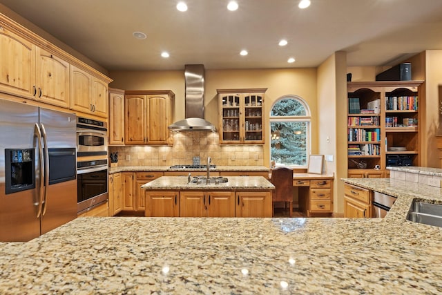 kitchen with wall chimney range hood, appliances with stainless steel finishes, a kitchen island with sink, built in desk, and light stone countertops