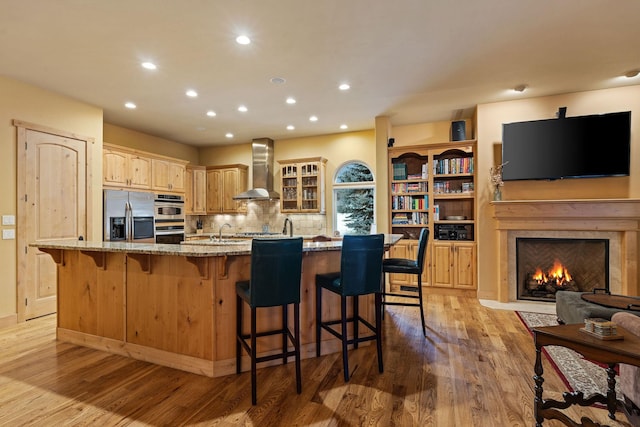 kitchen featuring a breakfast bar area, stainless steel appliances, a large island, light stone countertops, and wall chimney range hood