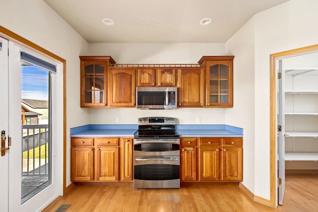 kitchen with brown cabinets, light wood-type flooring, glass insert cabinets, and stainless steel appliances