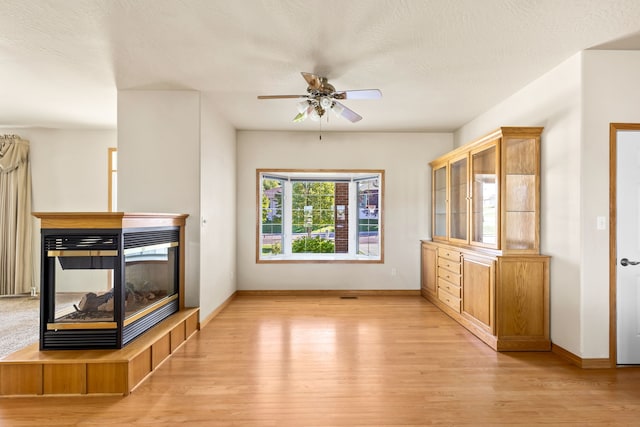 unfurnished living room featuring ceiling fan, a textured ceiling, a multi sided fireplace, baseboards, and light wood-style floors