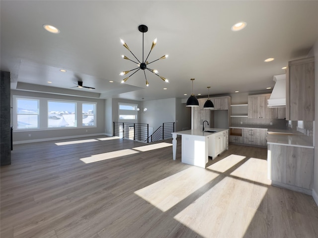 kitchen featuring tasteful backsplash, custom range hood, an island with sink, decorative light fixtures, and light wood-type flooring