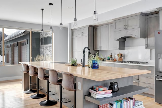 kitchen with pendant lighting, butcher block counters, a kitchen island with sink, stainless steel appliances, and light hardwood / wood-style flooring