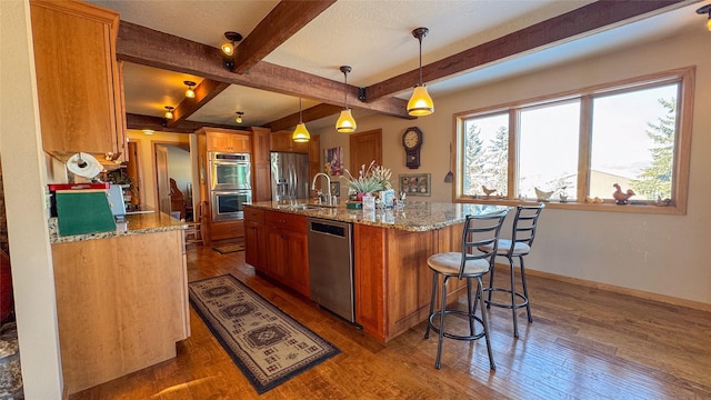 kitchen featuring appliances with stainless steel finishes, decorative light fixtures, beamed ceiling, an island with sink, and dark wood-type flooring