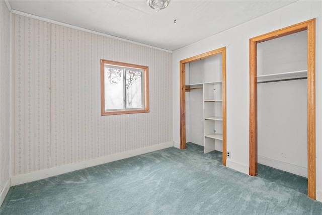 unfurnished bedroom with wallpapered walls, baseboards, dark colored carpet, crown molding, and multiple closets