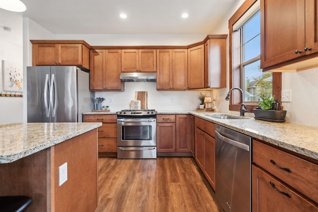 kitchen featuring appliances with stainless steel finishes, light stone counters, wood finished floors, under cabinet range hood, and a sink