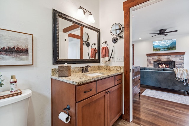 bathroom featuring toilet, ceiling fan, wood finished floors, a stone fireplace, and vanity