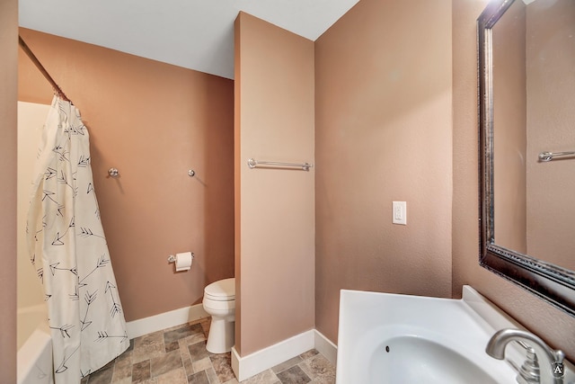 bathroom with shower / tub combo with curtain, toilet, stone finish flooring, a sink, and baseboards