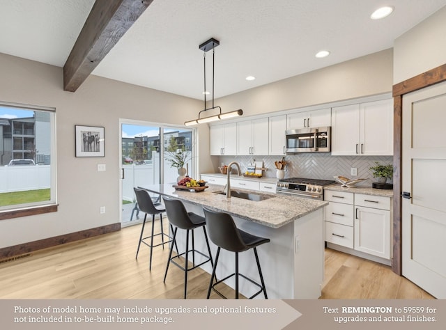 kitchen with decorative light fixtures, stainless steel appliances, a kitchen island with sink, a sink, and white cabinetry