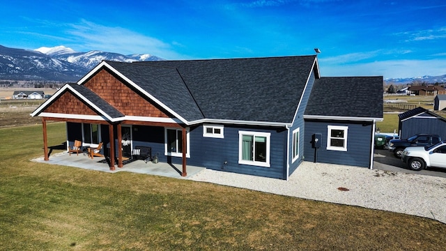 back of property with roof with shingles, a patio area, a mountain view, and a lawn