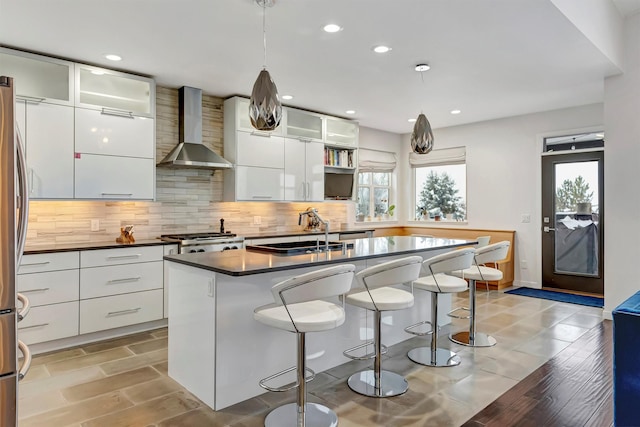 kitchen featuring white cabinets, dark countertops, glass insert cabinets, hanging light fixtures, and wall chimney range hood