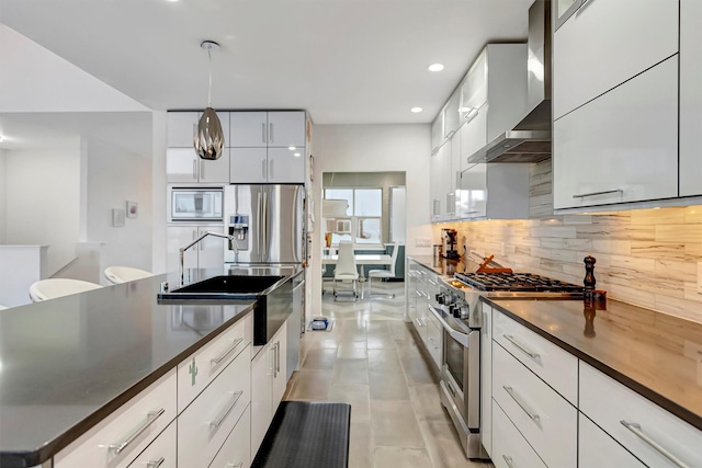 kitchen featuring white cabinets, dark countertops, stainless steel appliances, wall chimney range hood, and pendant lighting