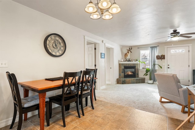 dining space featuring baseboards, light colored carpet, a tiled fireplace, and ceiling fan with notable chandelier