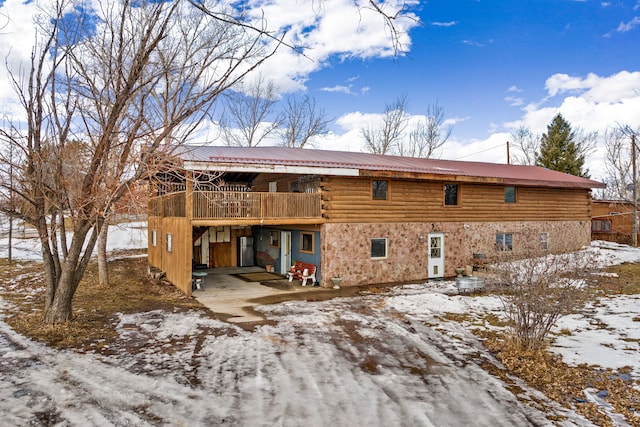 snow covered back of property featuring stone siding, metal roof, log exterior, and a balcony