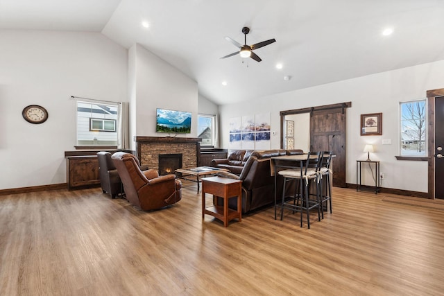 living room with a barn door, ceiling fan, a stone fireplace, light wood-style floors, and high vaulted ceiling