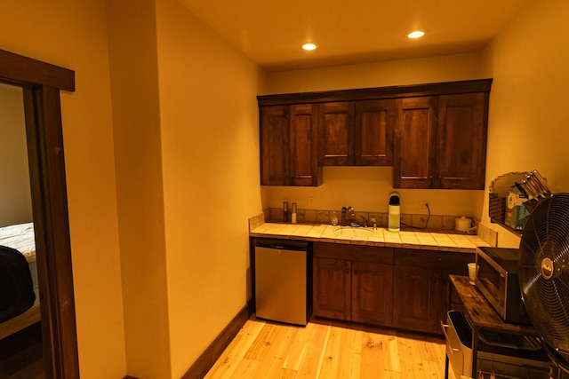 kitchen featuring tile counters, stainless steel appliances, dark brown cabinets, light wood-type flooring, and a sink