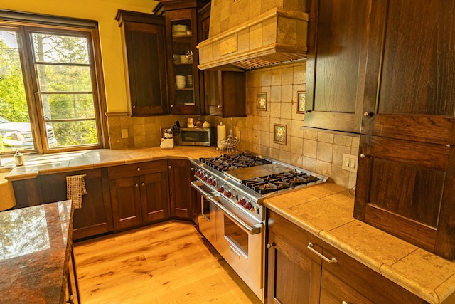 kitchen featuring decorative backsplash, light wood-style flooring, custom range hood, glass insert cabinets, and appliances with stainless steel finishes