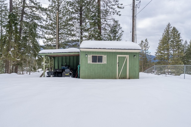 snow covered structure featuring a carport, an outdoor structure, and fence