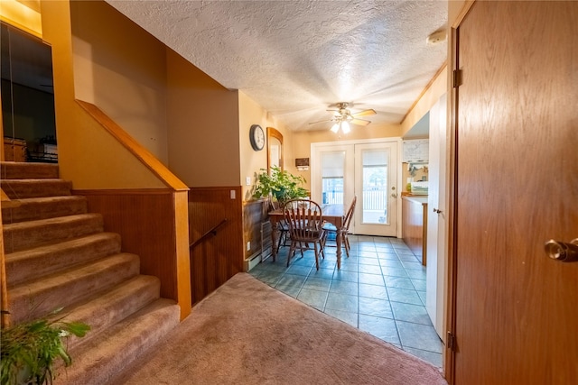 tiled dining space with a textured ceiling, ceiling fan, wooden walls, stairway, and wainscoting