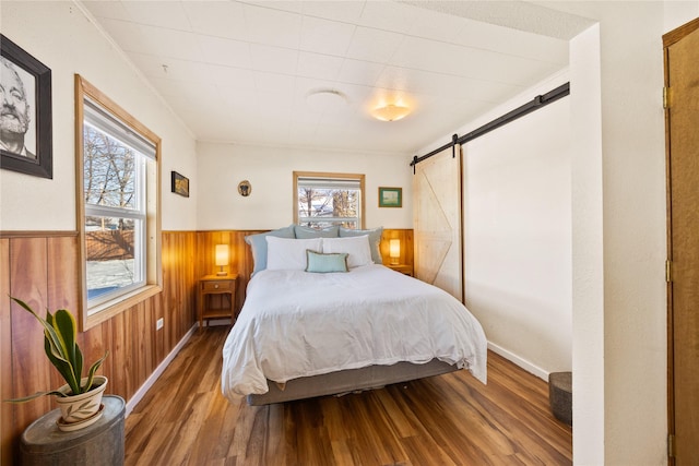 bedroom with a barn door, wooden walls, wood finished floors, ornamental molding, and wainscoting