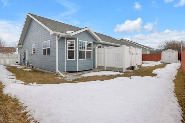 snow covered property featuring a shingled roof, an outbuilding, a fenced backyard, and a shed