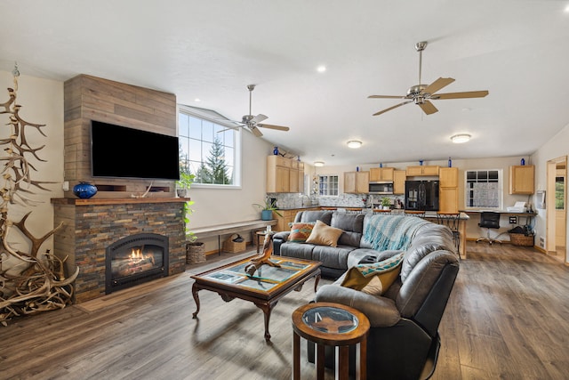 living room with a ceiling fan, vaulted ceiling, wood finished floors, and a stone fireplace