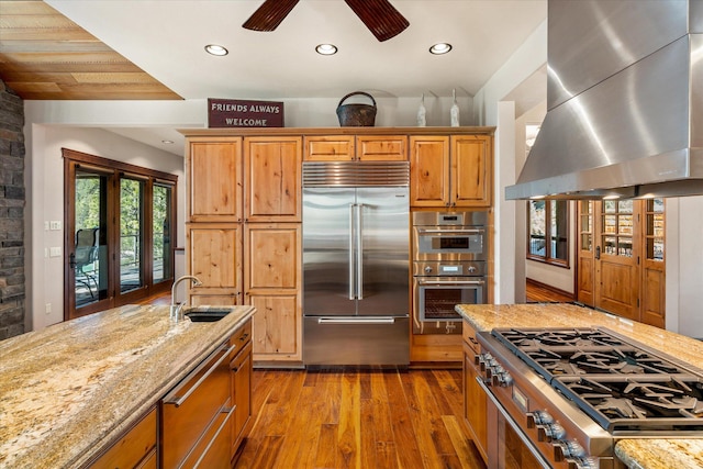 kitchen featuring light stone countertops, wall chimney range hood, stainless steel appliances, and a sink