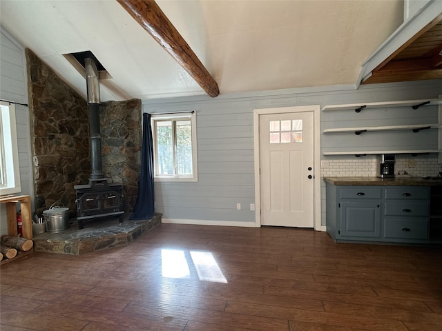 unfurnished living room with wooden walls, baseboards, lofted ceiling with beams, dark wood-type flooring, and a wood stove