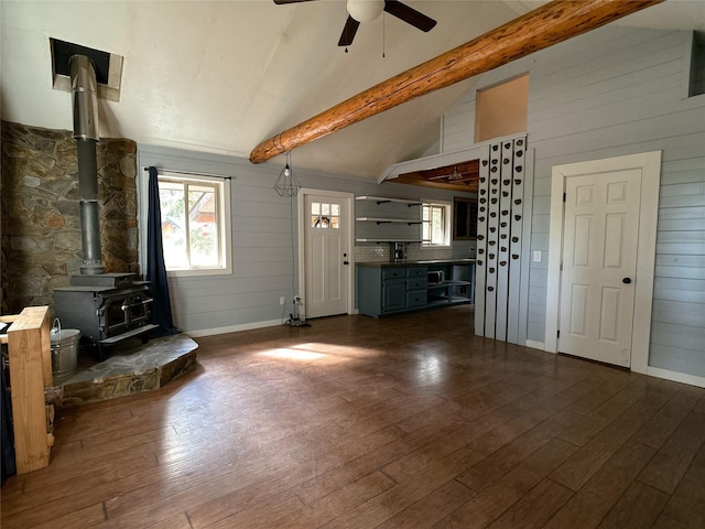 unfurnished living room with wooden walls, a ceiling fan, lofted ceiling with beams, dark wood-type flooring, and a wood stove