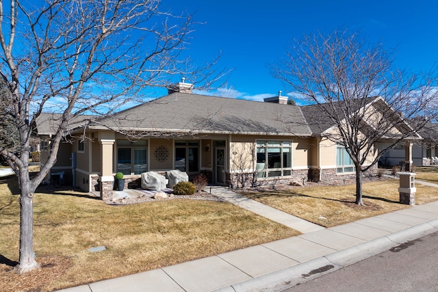 single story home featuring roof with shingles, a chimney, a porch, a front yard, and stone siding