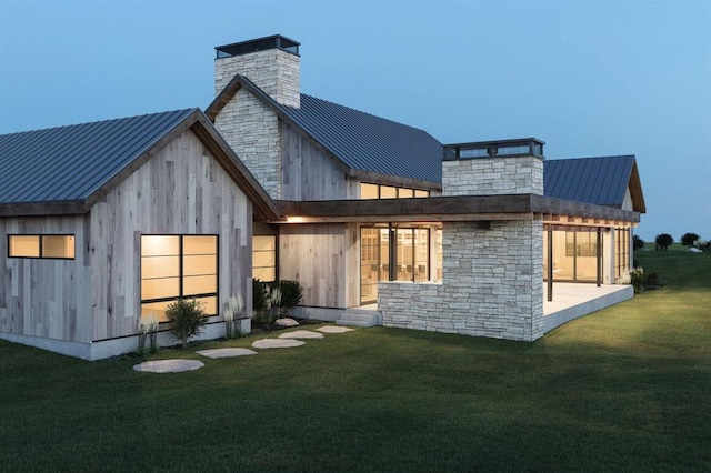 rear view of house featuring a lawn, stone siding, a chimney, metal roof, and a standing seam roof