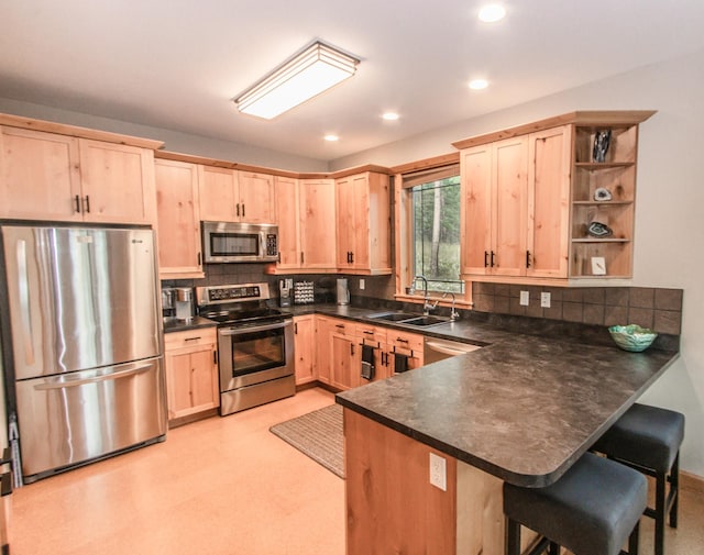 kitchen with dark countertops, appliances with stainless steel finishes, a peninsula, light brown cabinetry, and a sink